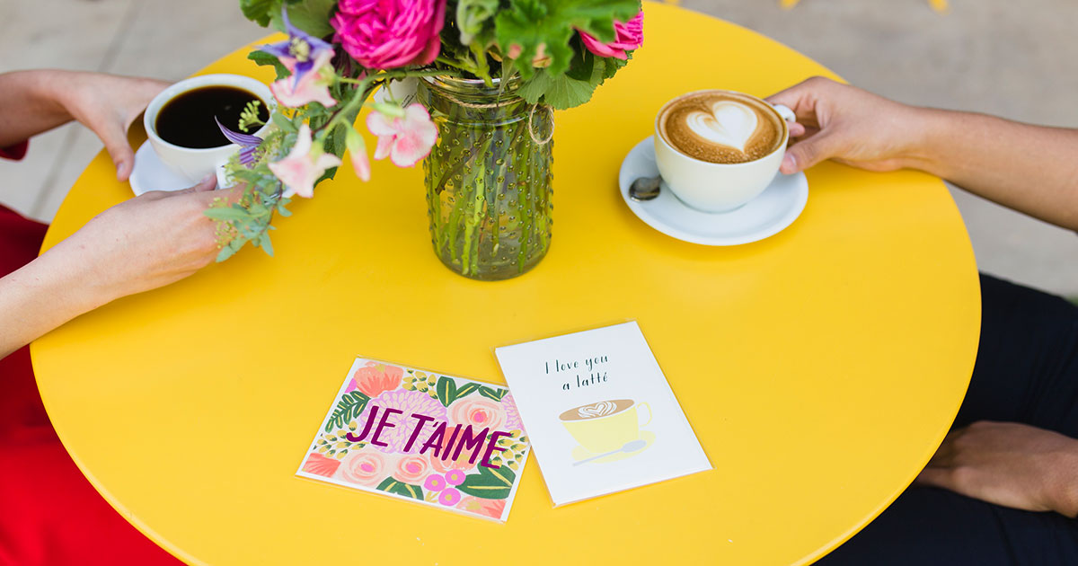 two people sitting opposite each other at a yellow table, each holding a cup of coffee, while two greeting cards sit between them
