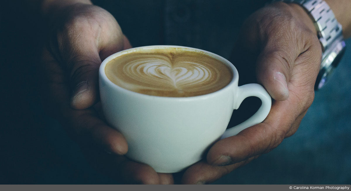 two hands holding a cappuccino in a white cup with heart latte art