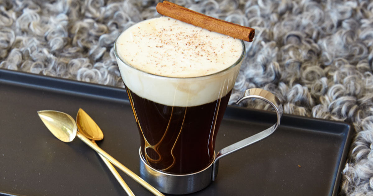 irish coffee in a clear glass with a large head of cream and a cinnamon stick resting on the edge
