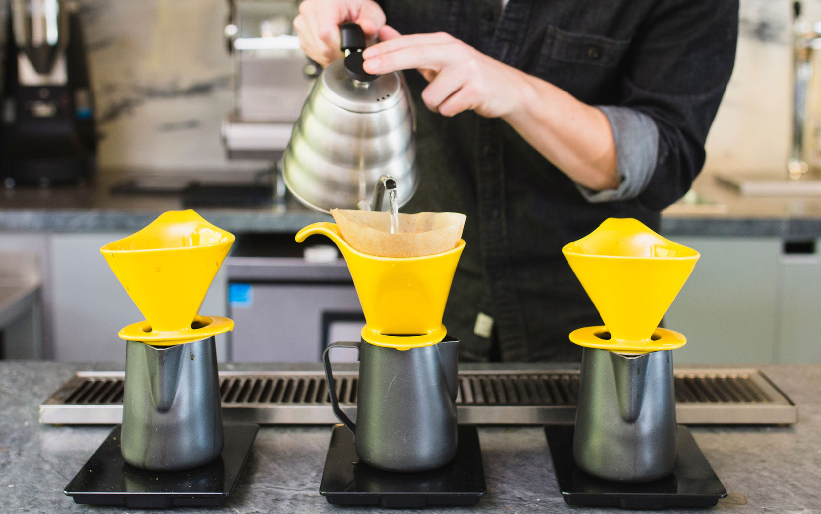 three sets of yellow bee house drippers, and black 20oz steaming pitchers sit in a row as a person brews coffee in the middle set, while pouring from a chrome hario v60 kettle