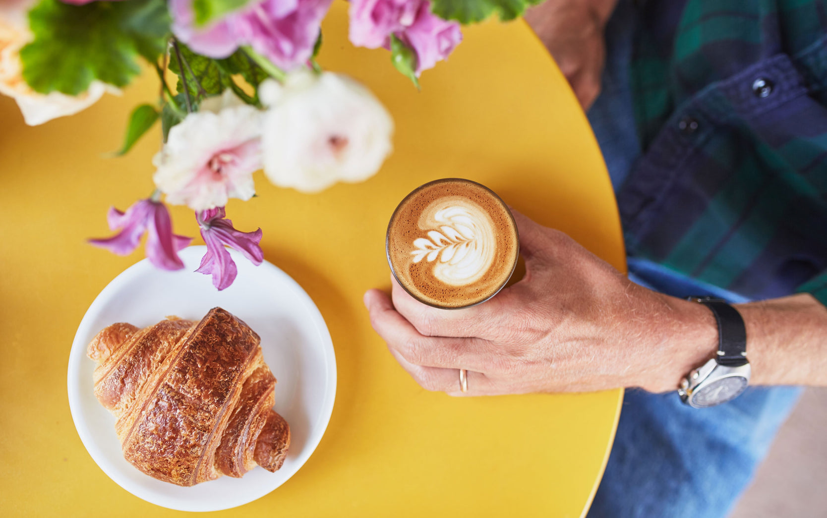 a person seated at a yellow table holding a gibraltar with rosetta latte art. a croissant on a plate sits at the table as well.