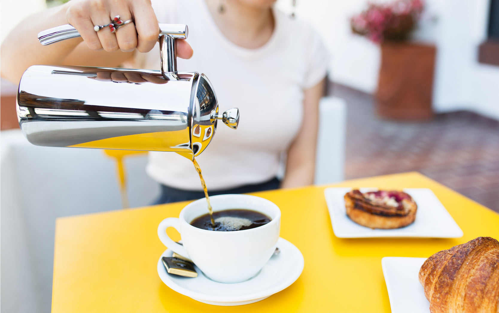 a person seated at a yellow table pouring coffee from a french press into a white coffee cup. there are also pastries on the table