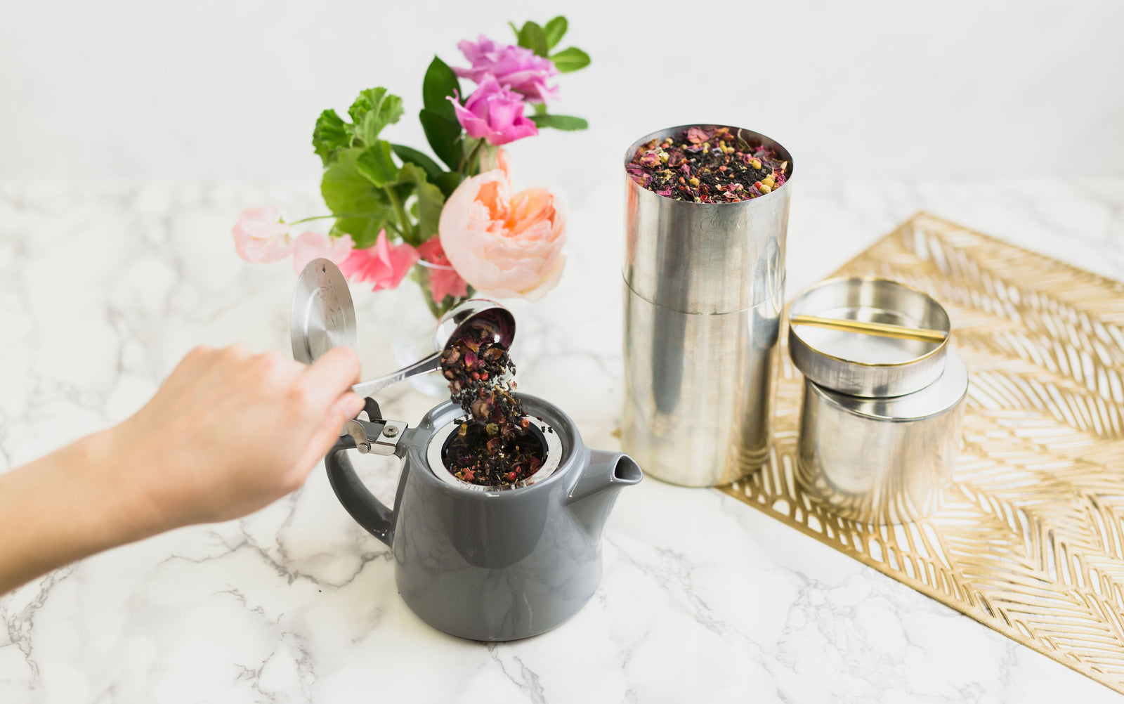 a hand pouring tea into a small grey tea pot, while a metal canister of said tea sits just above the pot.