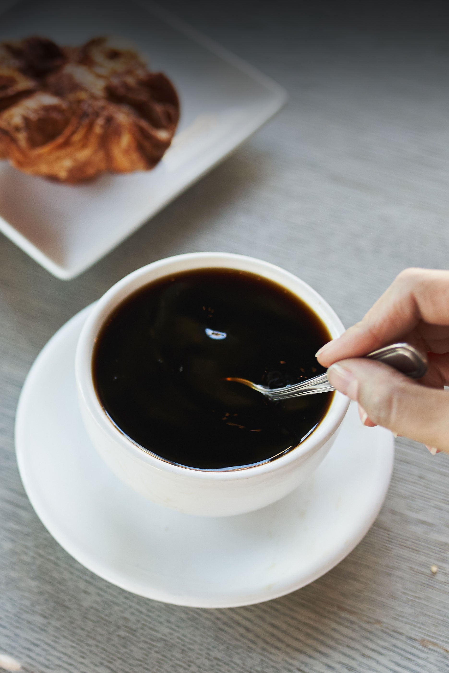 a cup of black coffee being stirred by a hand with a pastry in the frame