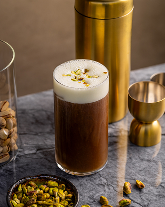Glass of iced coffee with a gold shaker and pistachios on a dark surface