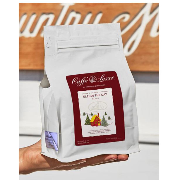 Caffè Lusso coffee bag labeled 'Sleigh the Day' held by a hand with a wooden background.